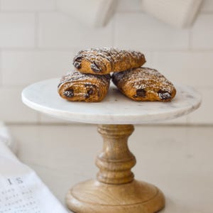 May include: Three chocolate croissants dusted with powdered sugar rest on a white marble cake stand with a wooden base. The stand sits on a light-colored countertop, with white mugs hanging in the background.