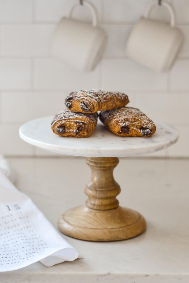 May include: Three chocolate croissants dusted with powdered sugar rest on a white marble cake stand with a wooden base. The stand sits on a light-colored countertop, with white mugs hanging in the background.