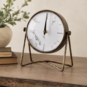 May include: A round table clock with a white face featuring a grey marble pattern and thin black markers. The clock is encased in a dark, distressed bronze-toned metal frame and is supported by a matching metal stand with a pivoting design.