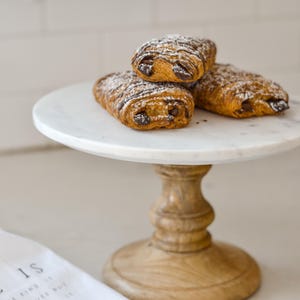 May include: Three chocolate croissants dusted with powdered sugar sit atop a white marble cake stand with a wooden base. The pastries are golden brown with visible layers and chocolate filling. A white cloth with text is in the foreground.