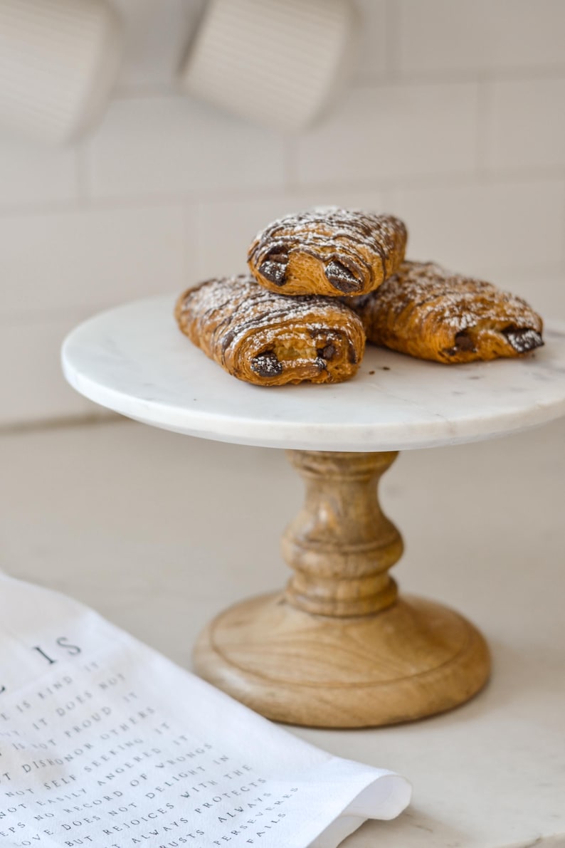 May include: Three chocolate croissants dusted with powdered sugar sit atop a white marble cake stand with a wooden base. The pastries are golden brown with visible layers and chocolate filling. A white cloth with text is in the foreground.