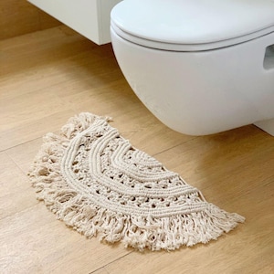 May include: A half-circle, cream-colored macrame rug with a fringed edge. The rug is placed on a light wood floor in front of a white toilet. The macrame design features concentric circles and open weave patterns.