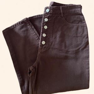 May include: Dark brown denim pants with a high waist and a button-up fly. The pants have a classic five-pocket design and a straight leg. The buttons are silver-colored.
