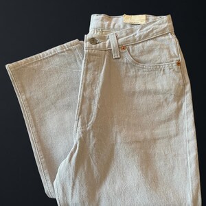May include: Light gray denim jeans with a classic five-pocket design. The jeans feature a button and zipper closure, along with copper rivets and a small tag. The jeans are folded, showcasing the front and side details.