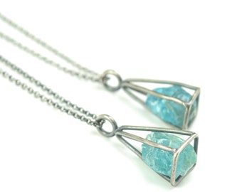 May include: Two silver necklaces with delicate chains and geometric pendants. Each pendant encloses a raw, light blue aquamarine stone.