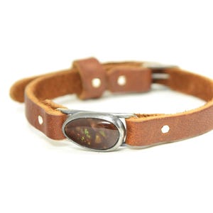 May include: A brown leather bracelet with a silver-toned oval centerpiece. The centerpiece features a brown and orange stone. The bracelet has a buckle closure and is set against a white background.