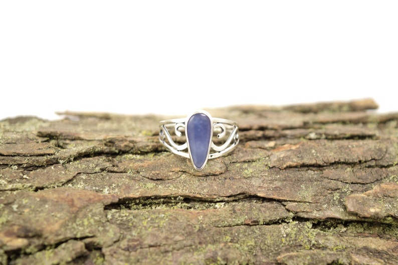 May include: A silver ring with a blue gemstone set in a teardrop shape. The ring is on a piece of tree bark.
