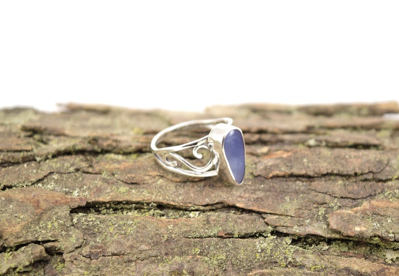 May include: A silver ring with a delicate filigree design and a purple gemstone. The ring is set on a piece of tree bark.