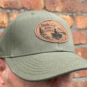 May include: An olive green baseball cap featuring an oval-shaped tan leather patch on the front. The patch displays a black engraved illustration of a person in a boat fishing on a lake with mountains and trees, and the text Dads gone fishing.