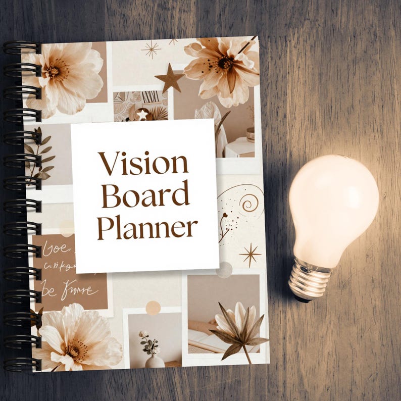 May include: A spiral-bound Vision Board Planner with a floral collage design in beige and brown tones. The planner has a white square overlay with the text "Vision Board Planner". A lit lightbulb is next to the planner on a wooden surface.