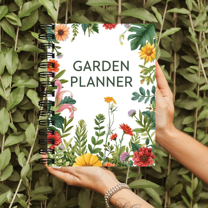 K&ouml;nnte beinhalten: Ein spiralgebundener Gartenplaner mit wei&szlig;em Einband, der farbenfrohe Blumenillustrationen und den Text "GARDEN PLANNER" in Gr&uuml;n zeigt. Der Planer wird vor einem Hintergrund aus gr&uuml;nem Laub gehalten.