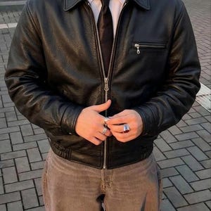 Op de afbeelding: Een zwart leren jack met een textuurafwerking, een zilverkleurige ritssluiting aan de voorzijde, een puntige kraag en een horizontale borstzak met rits. Het jack wordt gedragen over een wit overhemd met een zwarte das en een bruine broek.