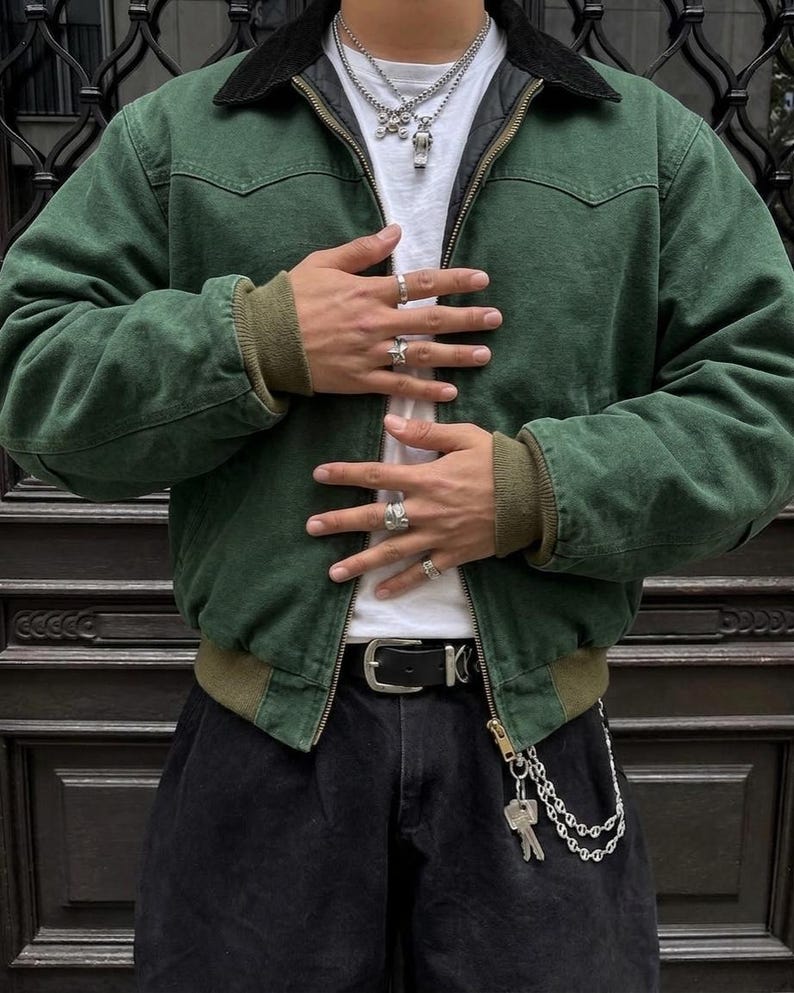 May include: A green jacket with a black collar, worn over a white t-shirt. The jacket has a zipper closure and ribbed cuffs. The person is wearing a black belt with a silver buckle and a chain with keys.