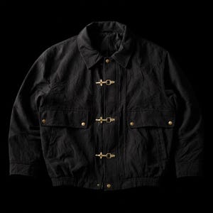 May include: Black jacket with a collared neckline, two front pockets, and gold-colored toggle closures. The jacket has a ribbed hem and cuffs. The fabric appears to be a textured, possibly cotton or a cotton blend.