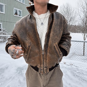 Könnte beinhalten: Eine braune Leder-Bomberjacke im Used-Look mit Stehkragen, Frontreißverschluss und gerippten Bündchen. Die Jacke wird über einem weißen Strickpullover mit Zopfmuster getragen und mit einer beigen Hose kombiniert. Eine silberne Kette ist sichtbar.