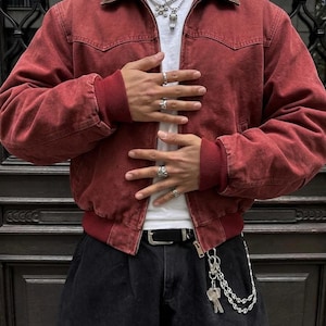 May include: A red canvas jacket with a brown corduroy collar, ribbed cuffs, and a ribbed hem. The jacket is worn over a white t-shirt, paired with black trousers featuring a black belt and a silver chain with keys attached to the belt loop.