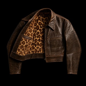 May include: A dark brown leather jacket featuring a pointed collar, long sleeves, and a front zipper closure. The interior lining is patterned with a tan and black leopard print, visible where the jacket is partially open at the front.