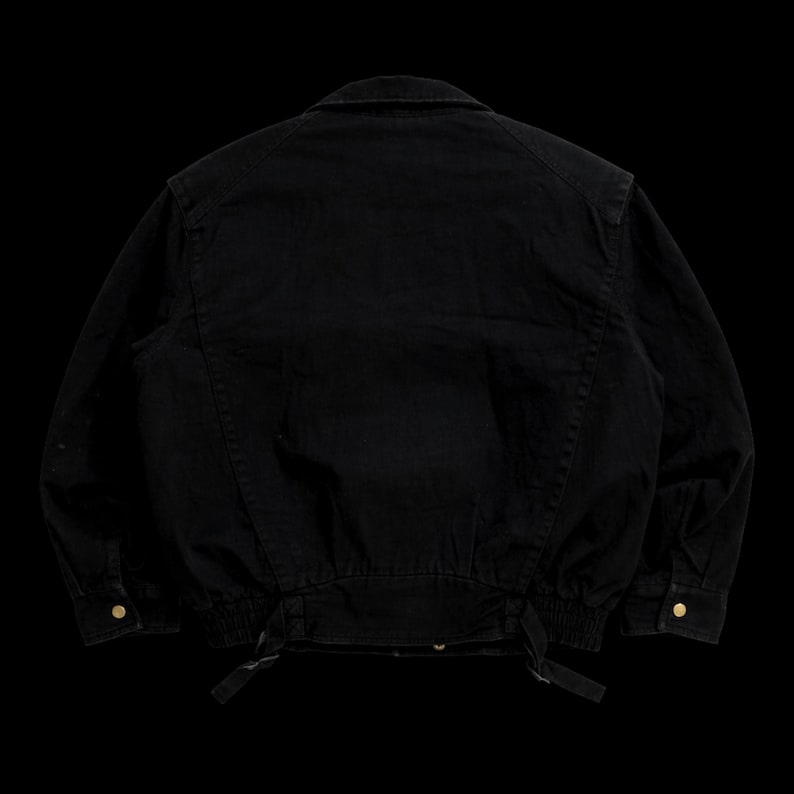 May include: A solid black denim jacket featuring a classic collar, long sleeves with buttoned cuffs, and a structured back panel. The hem includes an elasticized waistband with two adjustable fabric straps and metallic hardware accents.