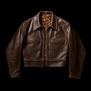 May include: A brown leather jacket with a classic collar and a zip-up front. The jacket features two chest pockets and a leopard print lining. The leather has a distressed appearance, giving it a vintage look. The jacket is displayed against a black background.