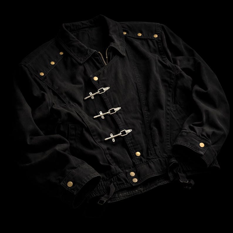 May include: A black jacket featuring a fold-down collar, long sleeves with buttoned cuffs, and a front closure with three silver-toned metal toggle clasps. The garment is accented with small gold-toned metal studs along the shoulders and hem.