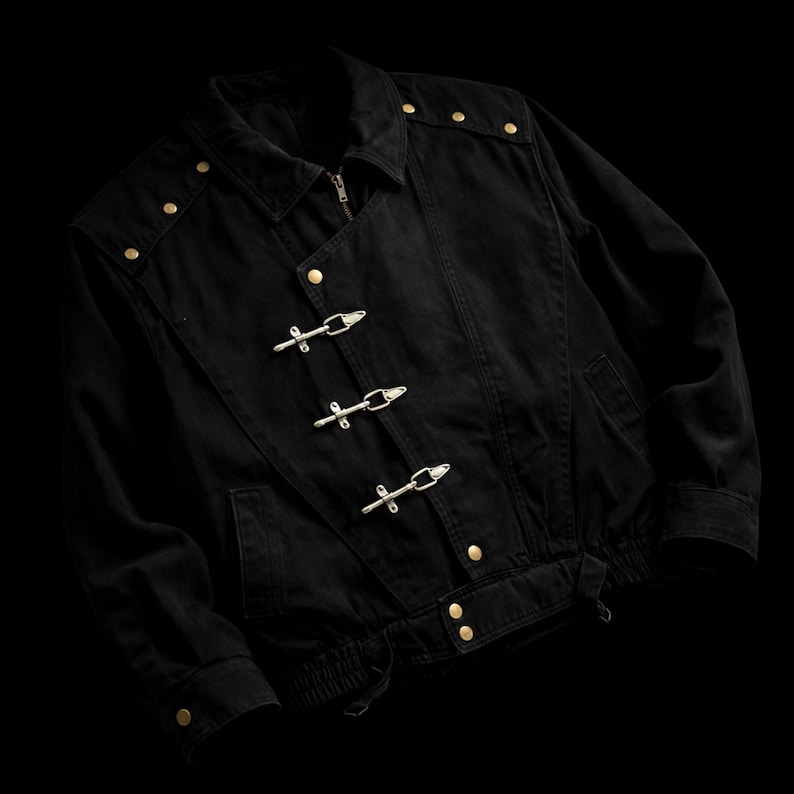 May include: A black jacket featuring a fold-down collar, long sleeves with buttoned cuffs, and a front closure with three silver-toned metal toggle clasps. The garment includes gold-toned metal buttons on the shoulders and waist hem.