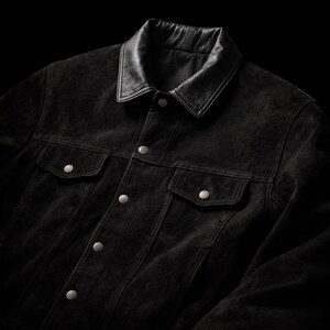 May include: A black suede jacket with a leather collar. The jacket features two chest pockets and two lower pockets, all secured with silver button closures. The jacket is fastened and has a classic design.