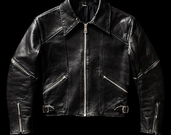 May include: A black leather jacket featuring a pointed collar, a full-length silver-tone front zipper, and two angled zippered chest pockets. The sleeves include zippered cuffs and upper arm zippers, while the waist has adjustable side buckles.
