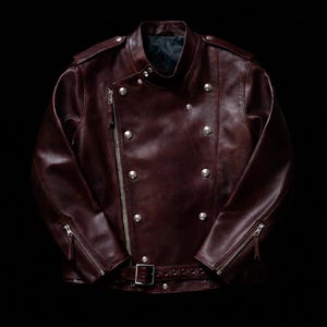 Può includere: Una giacca da moto in pelle color bordeaux scuro con chiusura asimmetrica a zip color argento, colletto alto con bottoni a pressione, molteplici bottoni in metallo argentato sul davanti, polsini con zip e cintura con fibbia in vita.