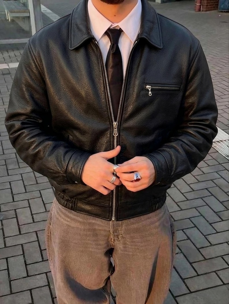 Op de afbeelding: Een zwart leren jack met een textuurafwerking, een zilverkleurige ritssluiting aan de voorzijde, een puntige kraag en een horizontale borstzak met rits. Het jack wordt gedragen over een wit overhemd met een zwarte das en een bruine broek.