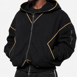 Pode incluir: Um casaco preto com capuz e fecho de correr, com debrum amarelo contrastante ao longo das costuras, um fecho frontal completo, punhos canelados e um pequeno bolso utilitário com fecho na manga esquerda. Peça com capuz.