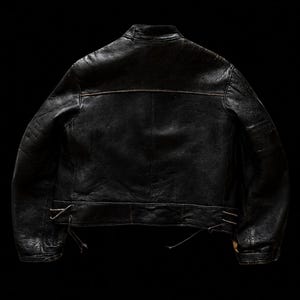 Puede incluir: Una chaqueta de cuero negro vista desde atr&aacute;s, con cuello alzado, mangas largas con pu&ntilde;os y una costura horizontal en la parte superior de la espalda. El dobladillo incluye cordones laterales ajustables con finas tiras de cuero colgando.