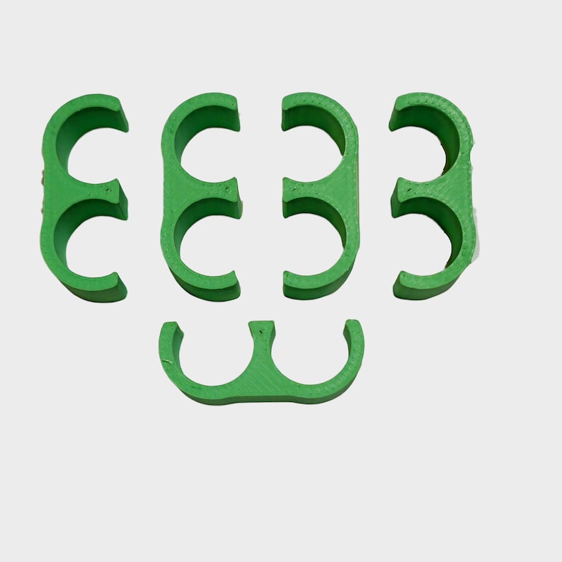 May include: A collection of six green plastic hose clamps, arranged in a scattered pattern against a white background. The clamps are semi-circular with a double-loop design, likely for securing hoses or pipes. The color is a vibrant, solid green.