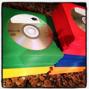 May include: A stack of green, red, blue, orange, and yellow DVD-R discs. The top disc has a graduation cap sticker and the text "VD+R".
