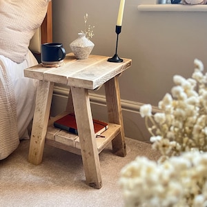 Op de afbeelding: Een kleine, houten bijzettafel met een onderste plank. Een donkerblauwe mok staat op een onderzetter, naast een kleine vaas en een zwarte kandelaar met een brandende kaars. Een rood notitieboekje ligt op de plank.