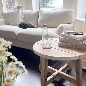 May include: A round wooden side table with a cross-base design, featuring a clear glass water bottle, a glass tumbler on a cork coaster, and a stack of three books titled "Life" and "Scandi Rustic" resting on its surface in a bright living room.