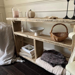 May include: A reclaimed wood console table with two shelves, featuring decorative items. A woven pumpkin, a ceramic bowl, a small basket, and books are on display. A grey blanket is draped on the side. Black candle holders and a glass cloche are on the top shelf.