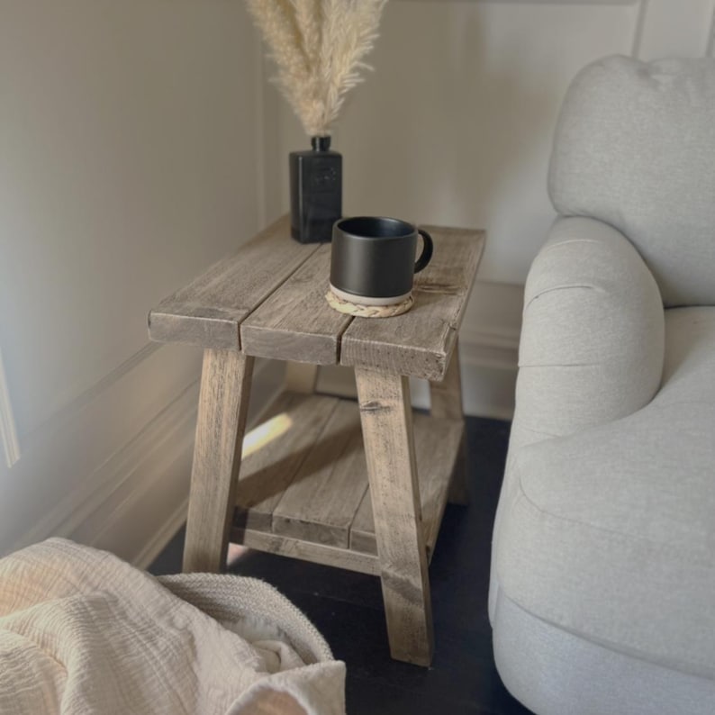 Op de afbeelding: Een rustieke, grijze houten bijzettafel met een onderste plank. Een zwarte mok staat op een geweven onderzetter op de tafel. Een zwarte vaas met gedroogd pampasgras staat er ook. Een lichtgekleurde deken ligt op de vloer.