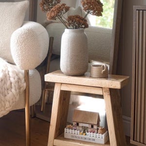 May include: A light-coloured wooden stool with a rectangular top and angled legs. On the top sits a speckled beige vase with dried brown flowers and a small ceramic mug. On the lower shelf is a wooden barn model with a white fence and text.