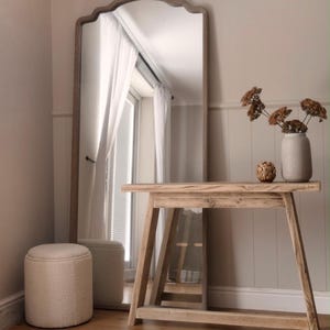 May include: A tall arched floor mirror with a wooden frame, a light-coloured wooden console table with angled legs, a cream-coloured textured cylindrical ottoman, and a light grey ceramic vase containing dried brown flowers on the table surface.