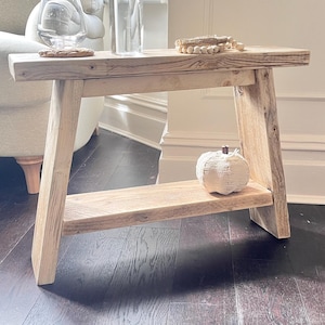 May include: A light-coloured wooden bench with a rectangular top and a lower shelf. The bench features angled legs and a visible wood grain texture. A small white decorative pumpkin sits on the lower shelf, while a glass and beads rest on top.
