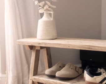 May include: A light-colored wooden bench with a lower shelf holding a pair of beige suede clogs and a pair of white and black sneakers. On the top surface sits a textured off-white ceramic jug containing a delicate branch with pale leaves.