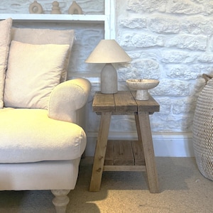 Op de afbeelding: Een woonkamer met neutrale tinten met een lichtgekleurde bank, een houten bijzettafel en een stenen lamp. De tafel heeft een lamp met een beige kap en een decoratieve kom. De achtergrond omvat een witte stenen muur en een plank met decoratieve objecten.