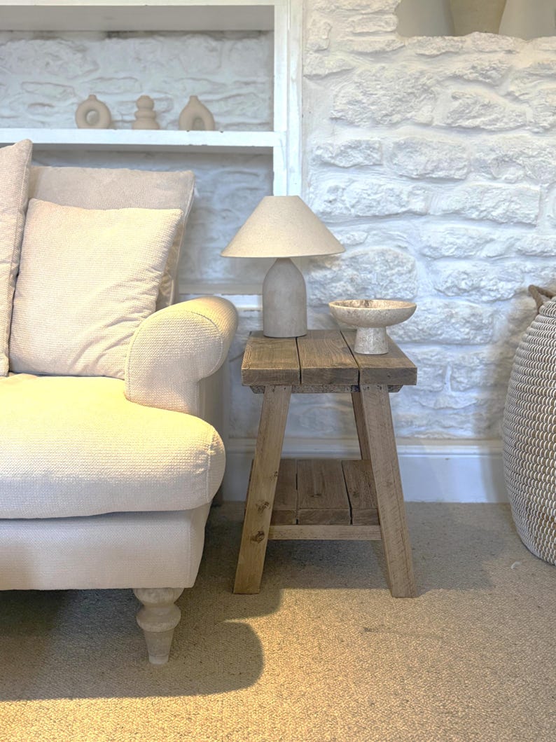 Op de afbeelding: Een woonkamer met neutrale tinten met een lichtgekleurde bank, een houten bijzettafel en een stenen lamp. De tafel heeft een lamp met een beige kap en een decoratieve kom. De achtergrond omvat een witte stenen muur en een plank met decoratieve objecten.
