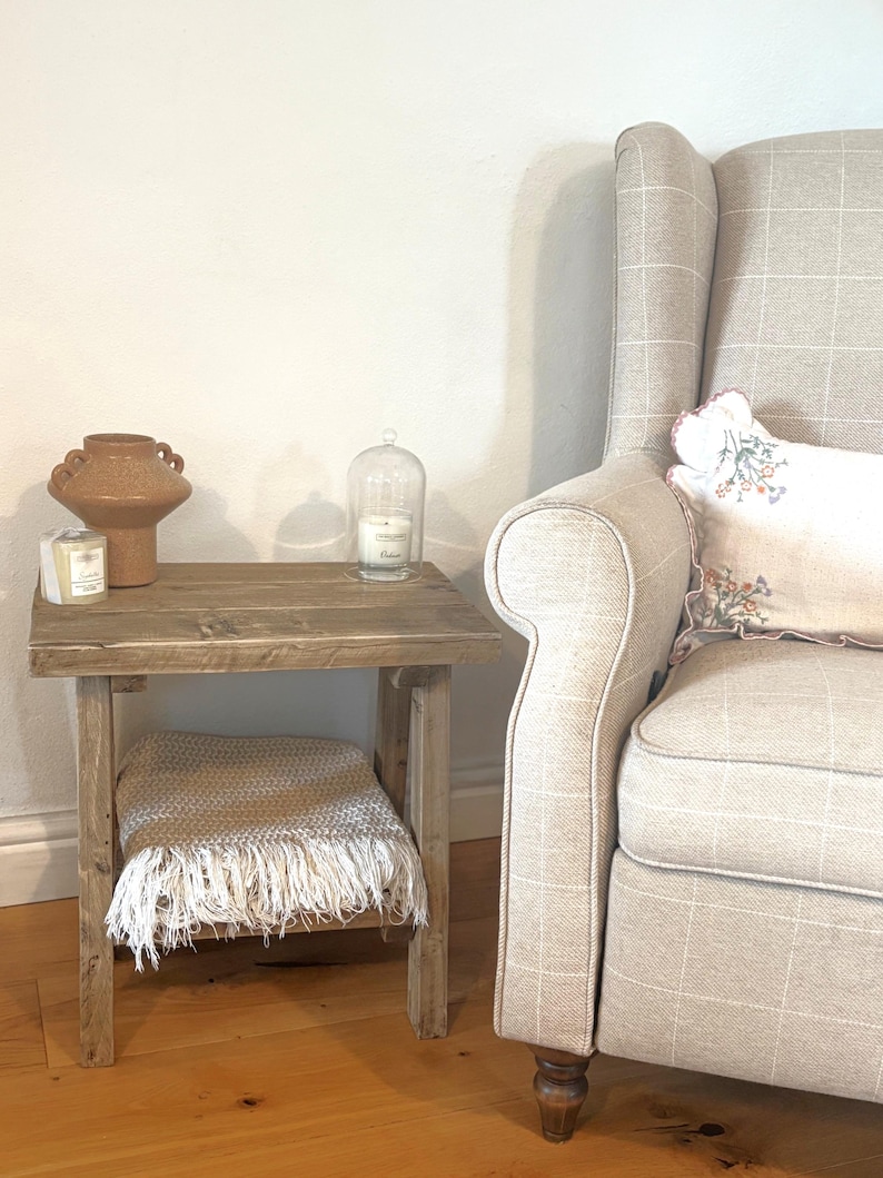 Op de afbeelding: Een rustieke houten bijzettafel met een gefranjerde deken op de plank. Een decoratieve vaas, een kaars en een kaars in een glazen stolp staan op het tafelblad. Rechts staat een fauteuil met patroon.