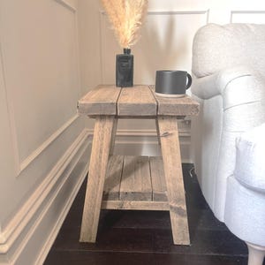 Op de afbeelding: Een rustieke houten bijzettafel met een plank. De tafel is gemaakt van verweerd hout en heeft een natuurlijke uitstraling. Een zwarte mok en een zwarte vaas met gedroogd pampasgras staan erop. De tafel staat naast een witte bank.