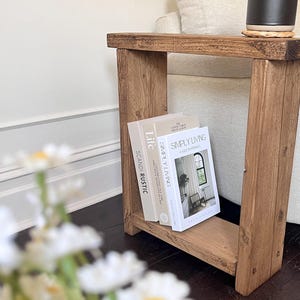 Könnte beinhalten: Ein rustikaler Beistelltisch aus Holz mit rechteckigem Design. Der Tisch hat ein unteres Regal mit drei Büchern mit weißen und beigen Einbänden. Auf der Oberseite steht ein schwarzer Becher neben einem beigen Untersetzer. Der Tisch ist aus hellbraunem Holz.