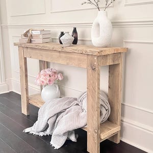 May include: A rectangular wooden console table with a natural light brown finish, featuring four straight legs and a lower shelf. The table surface holds books, small decorative vases, and a large white ceramic vase with a central cutout.