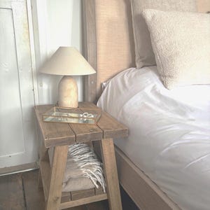 Op de afbeelding: Een rustieke houten bijzettafel met een beige lamp en een glazen dienblad. De tafel heeft een onderste plank met een opgevouwen deken. Op de achtergrond staat een bed met een beige hoofdbord en wit beddengoed.