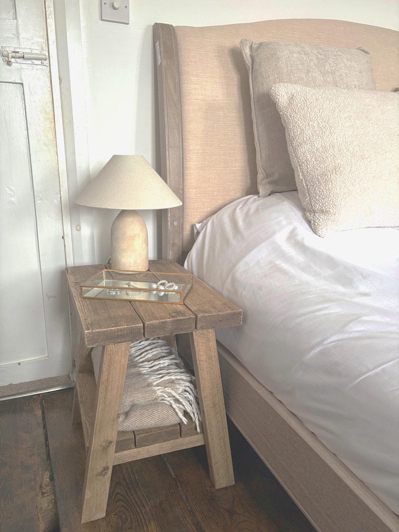 Op de afbeelding: Een rustieke houten bijzettafel met een beige lamp en een glazen dienblad. De tafel heeft een onderste plank met een opgevouwen deken. Op de achtergrond staat een bed met een beige hoofdbord en wit beddengoed.
