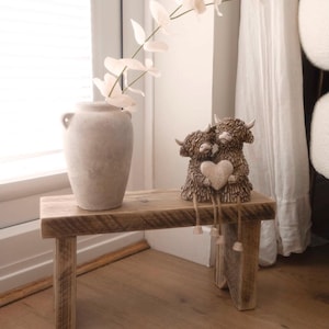 May include: A small rectangular wooden bench with a weathered finish. On top sits a light beige ceramic vase containing a delicate white floral stem, alongside a pair of brown Highland cow figurines holding a heart with dangling legs.